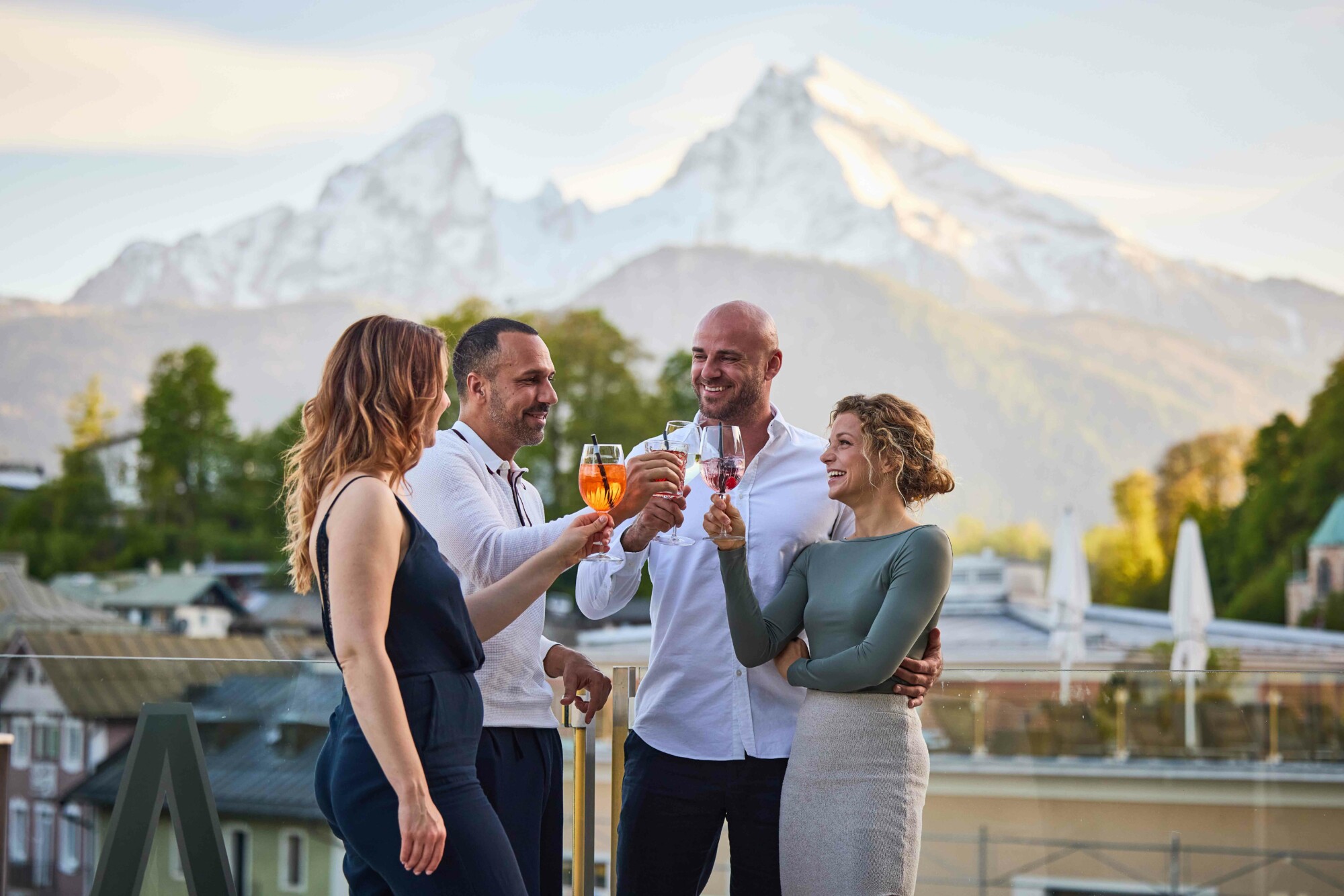 Vier Menschen, zwei Männer und zwei Frauen, feiern mit Gläsern Wein und Aperitif auf einem Balkon mit Blick auf Berge und Stadt im Hintergrund.