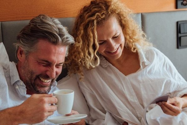 Ein Herr mit grauem Hemd und eine Dame mit weißem Oberteil lächelnd im Bett liegen mit einer Tasse Kaffee