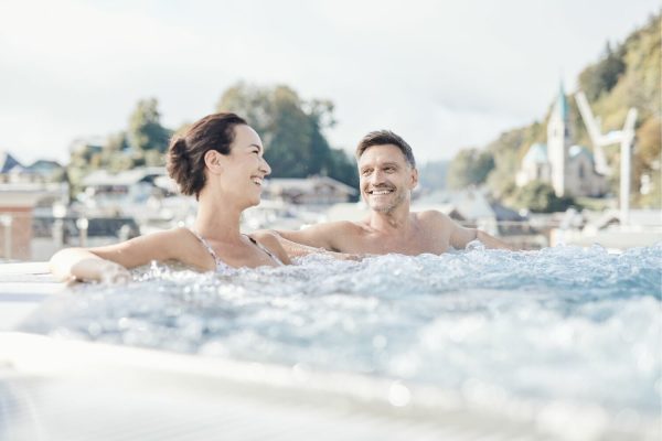 Ein lachendes Paar entspannt im Wasser auf einem Whirlpool mit einer malerischen Kulisse aus Häusern, Bäumen und einem Himmel im Hintergrund.