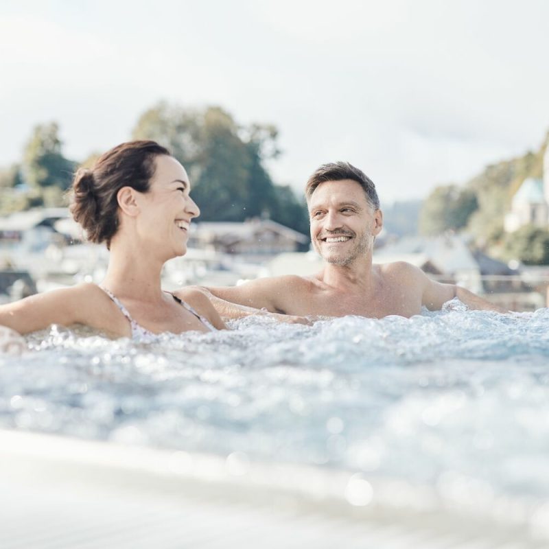 Ein lachendes Paar entspannt im Wasser auf einem Whirlpool mit einer malerischen Kulisse aus Häusern, Bäumen und einem Himmel im Hintergrund.
