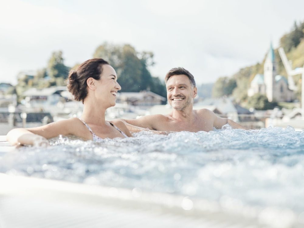 Ein lachendes Paar entspannt im Wasser auf einem Whirlpool mit einer malerischen Kulisse aus Häusern, Bäumen und einem Himmel im Hintergrund.