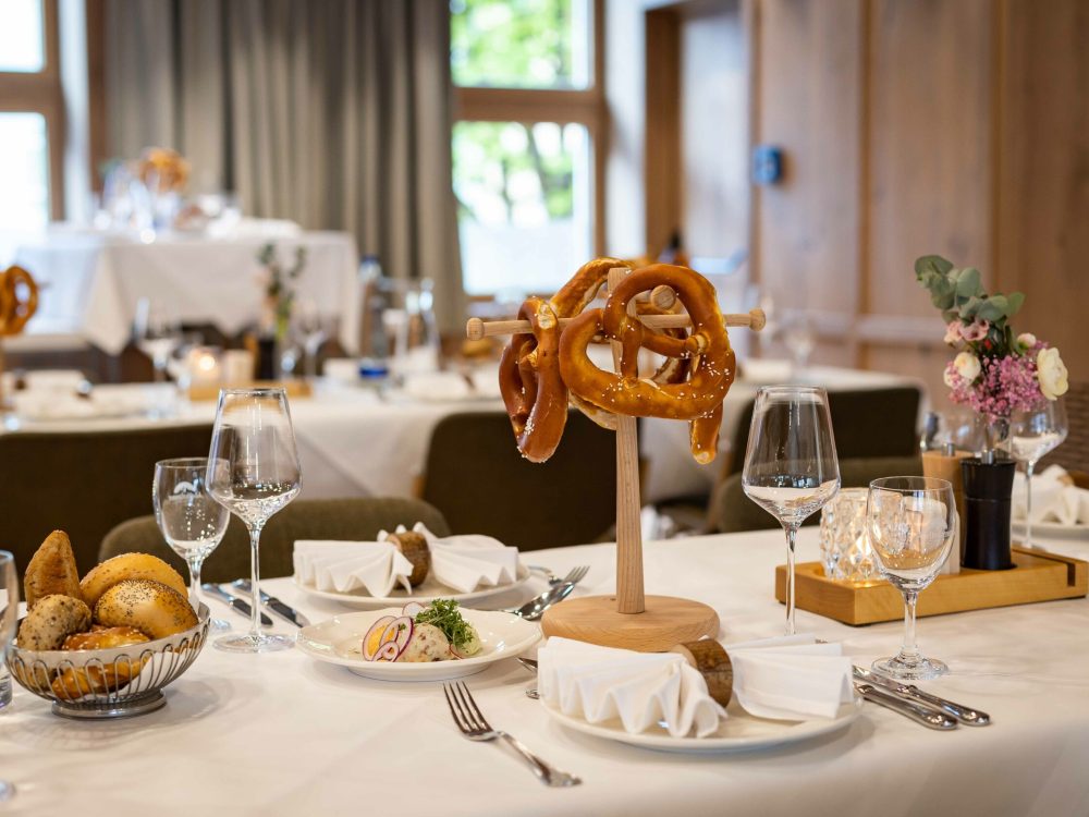 Ein festlich gedeckter Tisch mit einem zentralen Pretzel-Stand, umgeben von Weingläsern, Besteck, weißen Servietten und eleganten Dekorationen im Hintergrund.