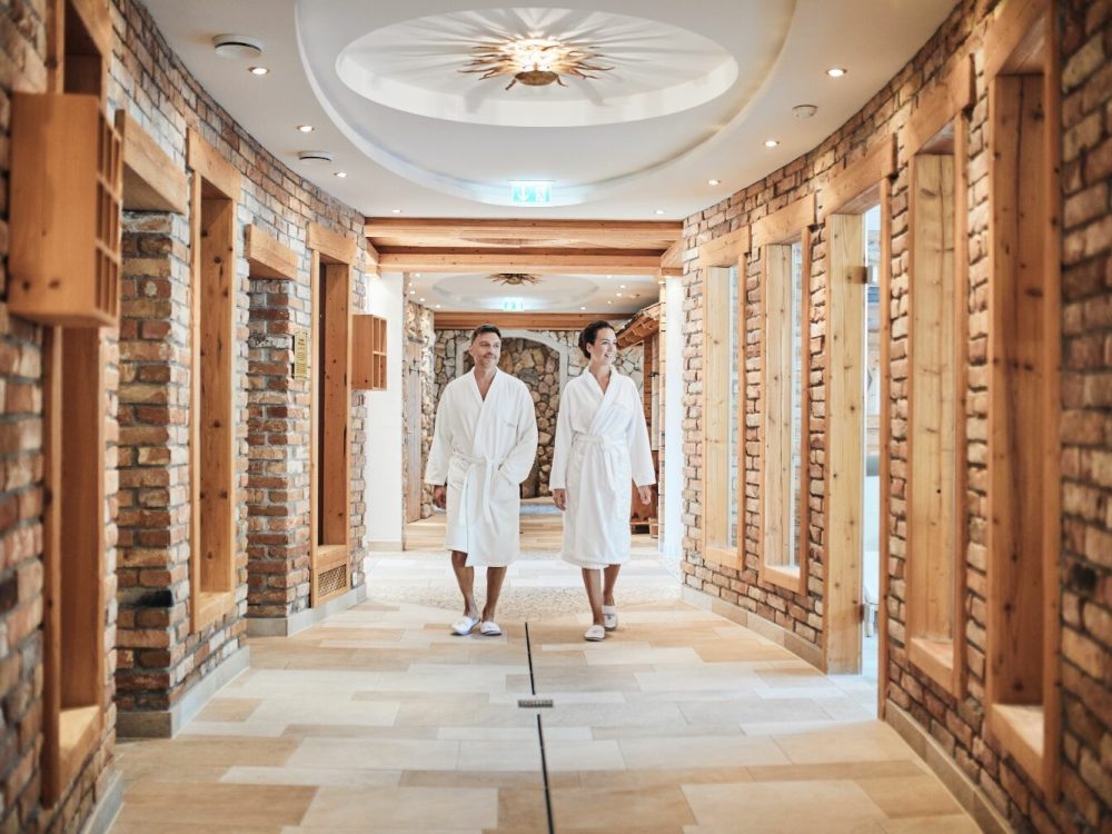 Zwei Personen in weißen Bademänteln gehen hand in hand durch einen gemütlichen Spa-Flur mit Ziegelwänden und Holzregalen.