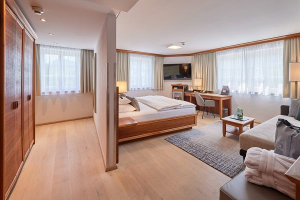 Ein Hotelzimmer mit einem großen Doppelbett, seitlichen Nachttischen mit Lampen, einem Schreibtisch mit Stuhl, einem Fernseher an der Wand, einem Sofa mit Kissen, großen Fenstern mit weißen und beigen Vorhängen und einer kleinen an den Fußende des Bettes stehenden Ablage.