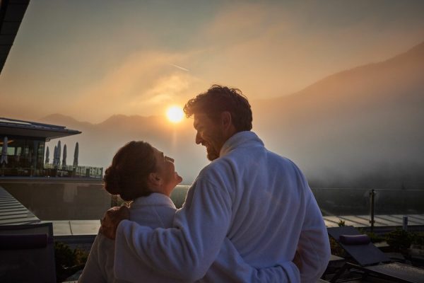 Ein Mann und eine Frau in Bademänteln auf einer Terrasse bei Sonnenuntergang, die sich liebevoll anschauen, mit Bergen im Hintergrund.