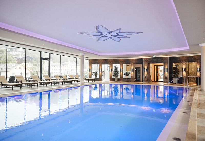 Innenansicht eines Schwimmbads mit rechteckigem Wasserbecken, gemütlichen Liegen am seitlichen Fenster, Pflanzen an der Wand und moderner Beleuchtung an der Decke.