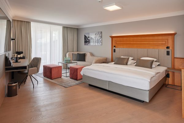 Schlafzimmer in einem Hotelzimmer mit einem großen Doppelbett, zwei Nachttischen, einem Sofa mit Kissen, einem kleinen Tisch mit Getränken, einem Schreibtisch mit Stuhl und einem Fernseher, im Hintergrund große Fenster mit Vorhängen.