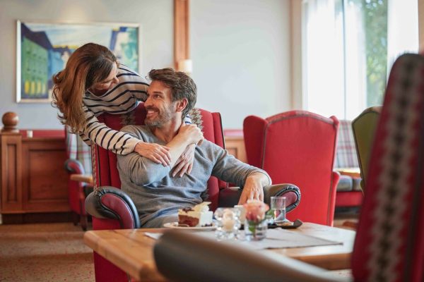Ein lachendes Paar im Wohnzimmer, der Mann sitzt im roten Sessel, die Frau lehnt sich zu ihm und umarmt ihn, während sie sich ansehen.