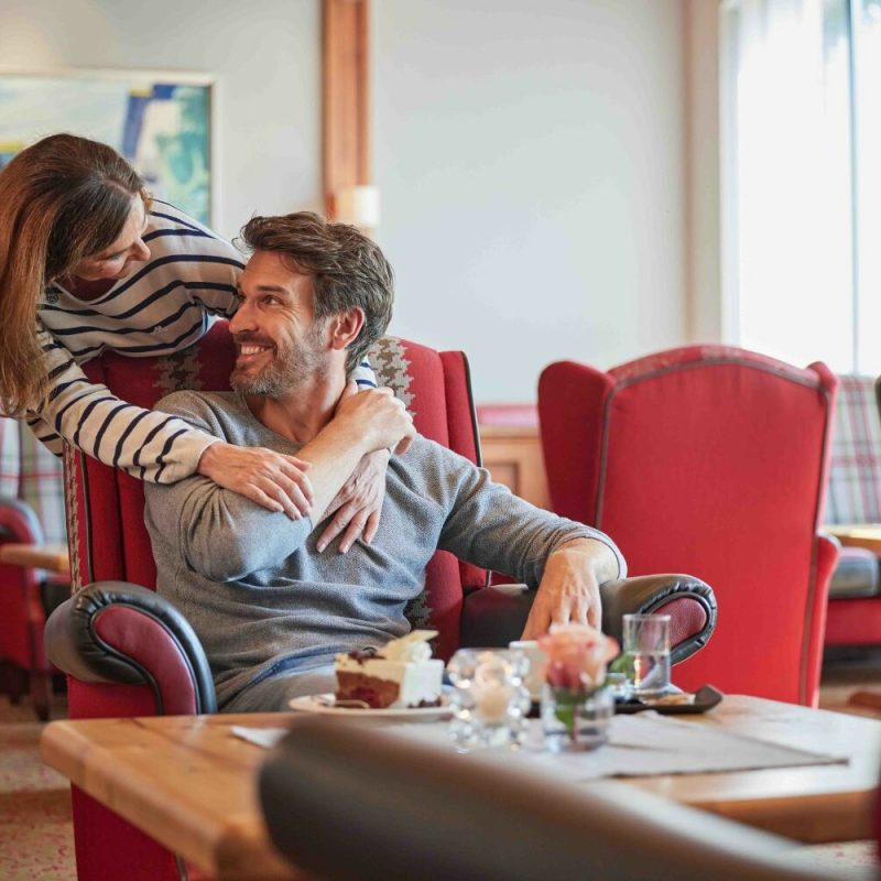 Ein lachendes Paar im Wohnzimmer, der Mann sitzt im roten Sessel, die Frau lehnt sich zu ihm und umarmt ihn, während sie sich ansehen.