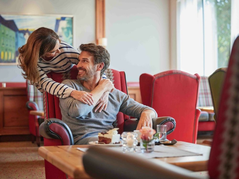 Ein lachendes Paar im Wohnzimmer, der Mann sitzt im roten Sessel, die Frau lehnt sich zu ihm und umarmt ihn, während sie sich ansehen.