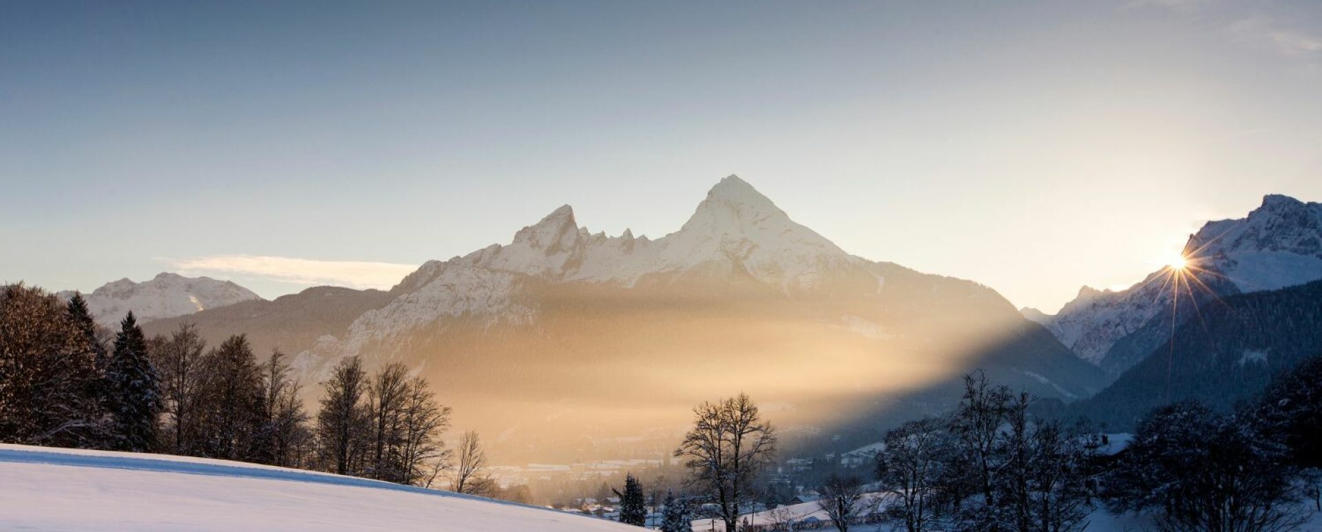 Schneebedecktes Tal mit vereinzelten Bäumen, im Hintergrund hohe, schneebedeckte Berge bei Sonnenaufgang, das Licht der Sonne blitzt zwischen den Bergen hervor.