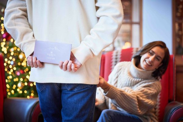 Eine Frau sitzt in einem gemütlichen Raum mit Weihnachtsdekoration, während eine andere Person in einem weißen Pullover und blauen Jeans ihr ein Geschenk überreicht, das in der Hand gehalten wird, wobei im Hintergrund ein geschmückter Weihnachtsbaum zu sehen ist.