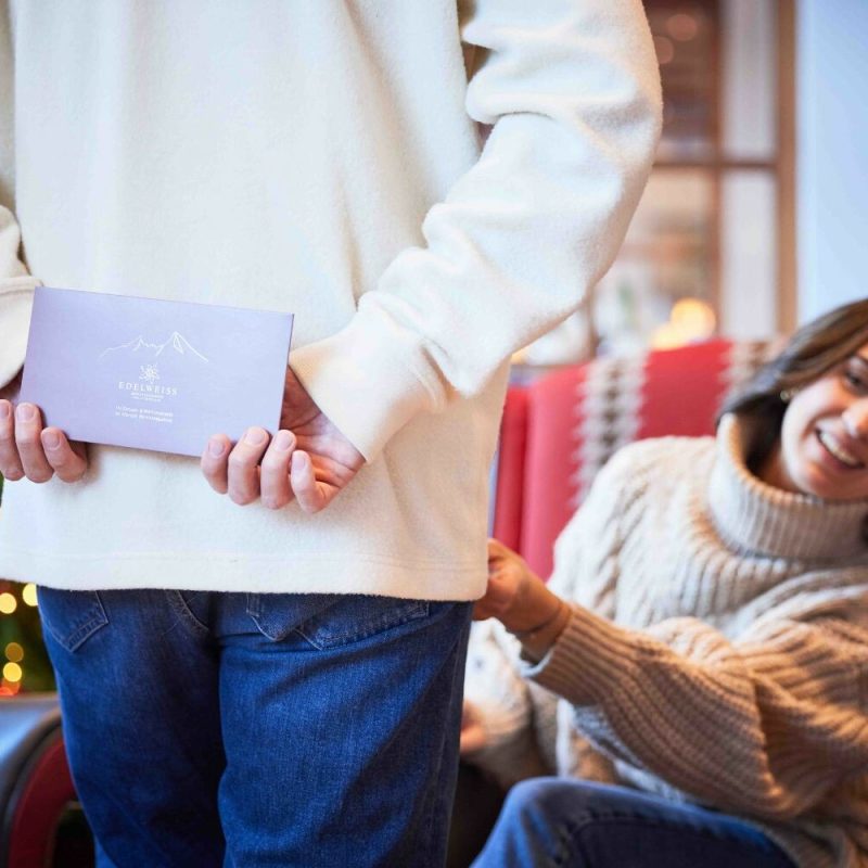 Eine Frau sitzt in einem gemütlichen Raum mit Weihnachtsdekoration, während eine andere Person in einem weißen Pullover und blauen Jeans ihr ein Geschenk überreicht, das in der Hand gehalten wird, wobei im Hintergrund ein geschmückter Weihnachtsbaum zu sehen ist.