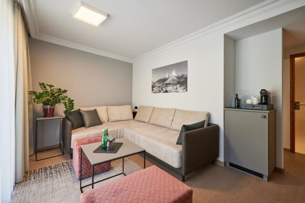 Wohnzimmer mit einer großen, beige Couch, schwarzen Kissen, einem Beistelltisch mit Flaschen und Gläsern, einem roten Ottomane, einem grauen Teppich und einer schwarzen und weißen Berglandschaftsfotografie an der Wand.