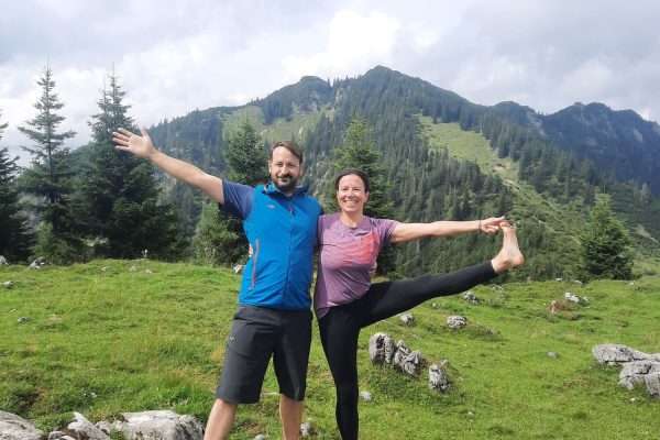 Ein Mann und eine Frau stehen nebeneinander auf einer grünen Wiese in den Bergen, die Frau macht eine Yogapose mit ausgestrecktem Arm und Bein, während der Mann lächelnd neben ihr steht, im Hintergrund sind grüne Hügel und Nadelbäume sowie bewölkter Himmel zu sehen.