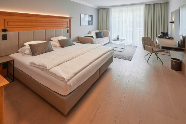 Ein Hotelzimmer mit Doppelbett, gepolstertem Kopfteil, Nachttischen, einem beige Sofa mit Kissen, einem kleinen Couchtisch mit Wasserflaschen, einem Schreibtisch mit Stuhl, einem Fernseher an der Wand und großen Fenstern mit weißen Vorhängen.