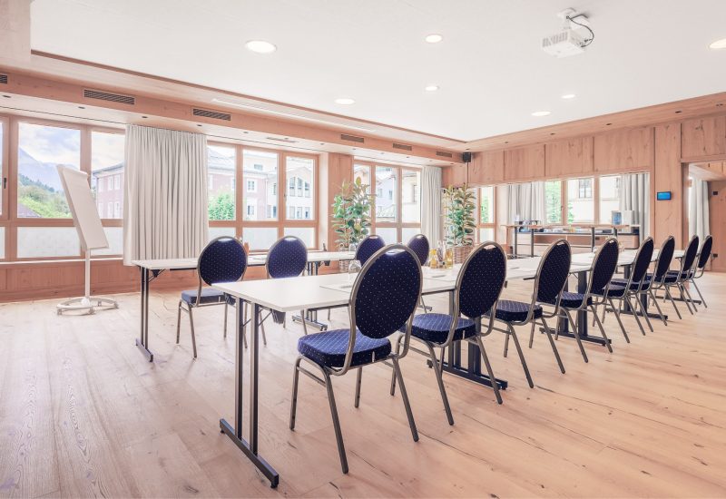 Ein helles, modernes Konferenzzimmer mit großen Fenstern, weißen Tischen, schwarzen Stühlen mit gepunktetem Bezug, Pflanzen und einem Flipchart auf einem Ständer.
