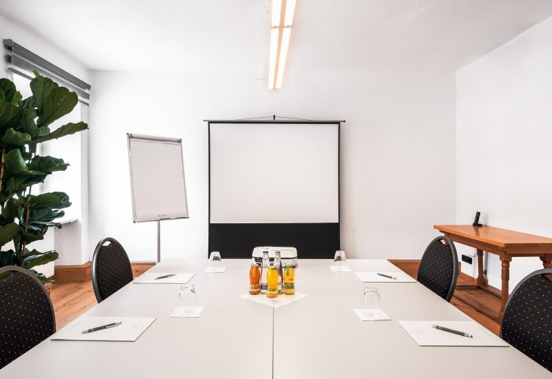 Konferenzraum mit weißem Tisch, sechs schwarzen Stühlen mit goldenen Punkten, Wasserflaschen und Gläsern, ein Whiteboard, eine Leinwand, eine kleine Holztisch und ein großes Pflanz in der Ecke.