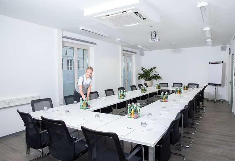 Ein gut beleuchteter Konferenzraum mit weißen Tischen und schwarzen Stühlen, zwei große Fenster, eine Flipchart, und eine Frau beim Platzdecken, bereit für ein Meeting.