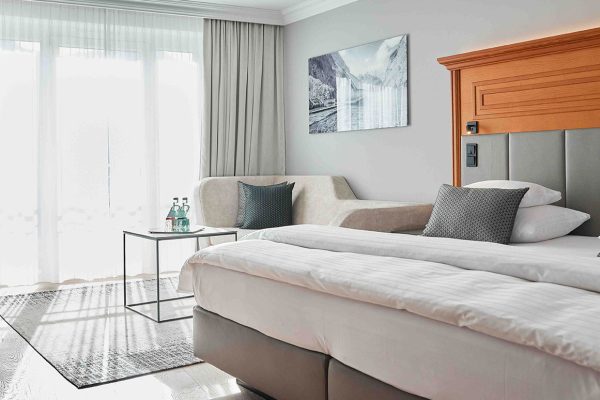 Ein Hotelzimmer mit einem Doppelbett, grauem Kopfteil und grauen Kissen, einem kleinen Tisch mit Wasserflaschen, einem Sofa mit Kissen, großen Fenstern mit weißen Vorhängen und einer Wanddekoration mit Berg- und Wasserfallmotiv.