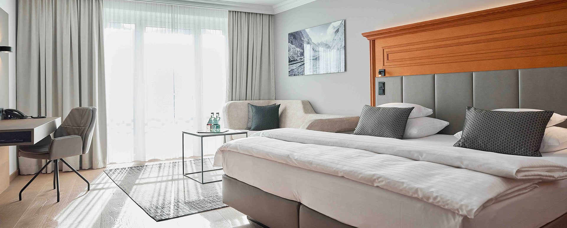 Ein Hotelzimmer mit einem Doppelbett, grauem Kopfteil und grauen Kissen, einem kleinen Tisch mit Wasserflaschen, einem Sofa mit Kissen, großen Fenstern mit weißen Vorhängen und einer Wanddekoration mit Berg- und Wasserfallmotiv.