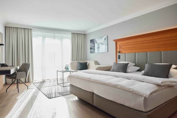 Ein modernes Hotelzimmer mit einem Doppelbett, grauem Polsterkopfteil, vier dunklen Kissen, einem beige-grauen Sofa mit Kissen, einem schwarzen Beistelltisch mit Wasserflaschen, einem Schreibtisch mit Computer und Telefon, einem großen Fenster mit weißen Vorhängen und einer Wandkunst mit Berglandschaft.