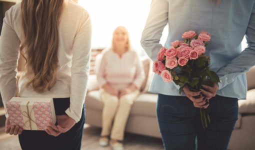 Frau mit langem, braunem Haar hält ein Geschenk, Frau mit blauer Bluse hält einen Blumenstrauß, im Hintergrund sitzende ältere Frau im weißen Hemd und bequemer Kleidung.