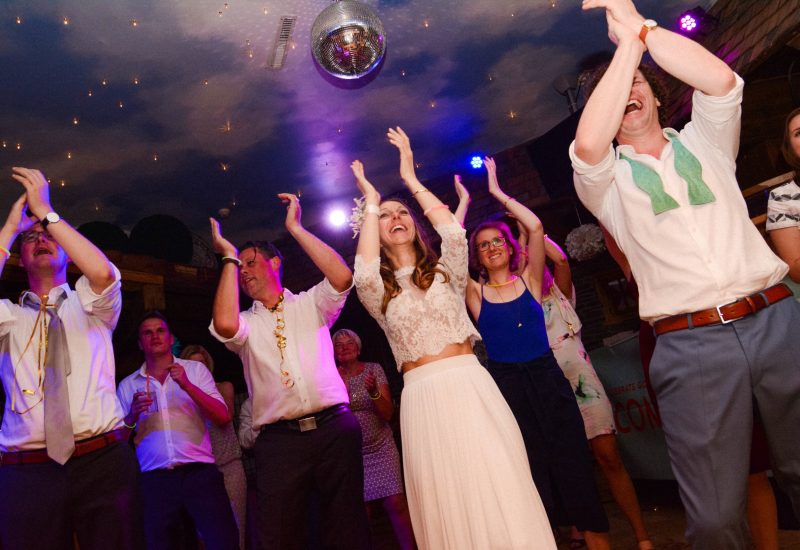 Menschen, die bei einer Feier auf der Tanzfläche tanzen, mit einem Disco-Ball an der Decke und bunten Lichtern im Hintergrund.