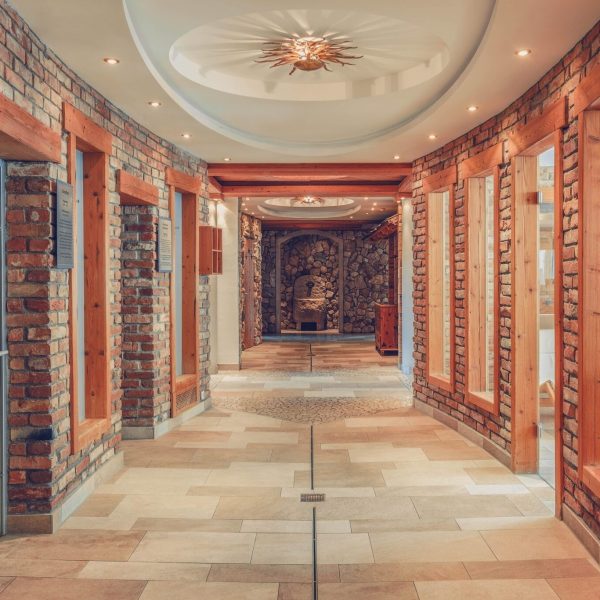 Ein heller Flur mit Wänden aus roten Ziegelsteinen und dekorativen Holzrahmen, mit großen Fensterflächen auf der rechten Seite und einer Feuerstelle oder einen Wasserbrunnen im Hintergrund, Holzfußboden und eine zentrumsliegende Bodenmarkierung.