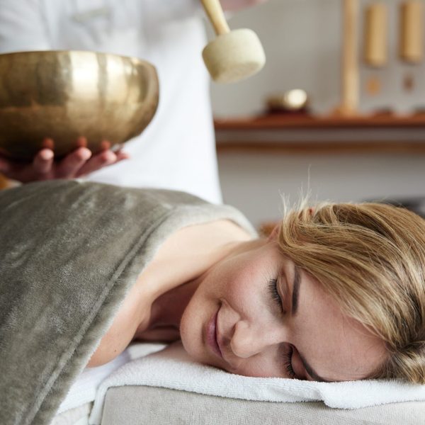 eine Frau liegt entspannt mit geschlossenen Augen auf einer Liege in einer Massagekabine, während eine Masseurin im Hintergrund mit einem Mallet auf eine Klangschale schlägt