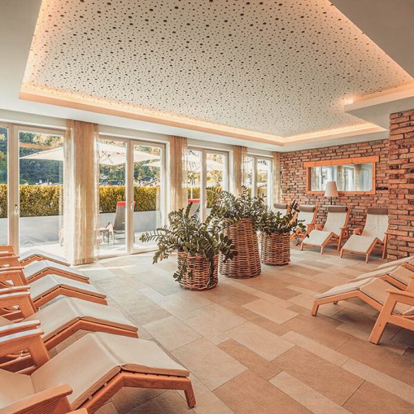 Ein Sonnenraum oder Spa-Bereich mit Sandsteinboden, braunen Ziegelwänden, großen Fenstern mit leichten Vorhängen, mehreren Holzliegen mit weißen Kissen, zentralen Pflanzen in Körben, und einem dekorativen abgehängten Deckenbereich mit Beleuchtung.