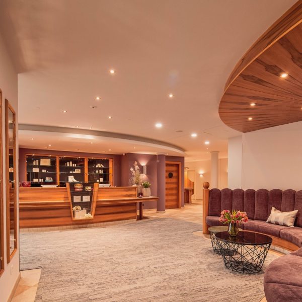 Ein elegantes Hotel-Lobby mit einem hohen, runden Empfangstresen aus Holz und einer Bücherwand im Hintergrund, einer halbkreisförmigen, gepolsterten Sitzecke mit Kissen und einem kleinen Tisch mit Blumen, moderne Wand- und Deckenleuchten sowie eine helle, einladende Atmosphäre.