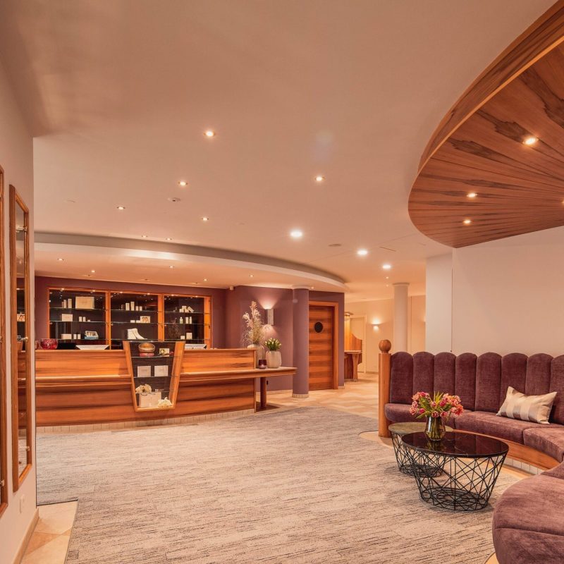 Ein elegantes Hotel-Lobby mit einem hohen, runden Empfangstresen aus Holz und einer Bücherwand im Hintergrund, einer halbkreisförmigen, gepolsterten Sitzecke mit Kissen und einem kleinen Tisch mit Blumen, moderne Wand- und Deckenleuchten sowie eine helle, einladende Atmosphäre.