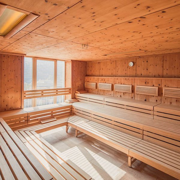 Ein Spa- oder Saunaraum mit Holzwänden, Decke und lange Bänke entlang der Wände, große Fenster, die natürliches Licht hereinlassen, und eine kleine Teppichmatte auf dem Boden.