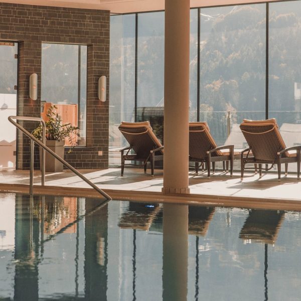 Innenbereich eines Spa- oder Hotelpools mit sechs Liegen, große Fenster mit Blick auf eine Berglandschaft, Pflanzen in Pflanzkübeln und ein dunkles Ziegelmauer-Design im Hintergrund.