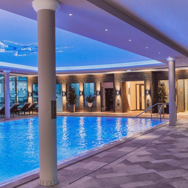 Innenaufnahme eines schwimmenden Pools in einer modernen Wellness-Oase, umgeben von Liegen, großen Fenstern und Pflanzen, mit blauer Beleuchtung und einer abendlichen Stadtansicht im Hintergrund