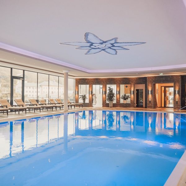 Innenaufnahme eines schwimmbads mit blauer Wasseroberfläche, umgeben von Liegestühlen und großen Fenstern, die den Blick auf eine Stadt mit Hügeln im Hintergrund bieten, und einer Deckenlila-Umrahmung mit dekorativem Licht.