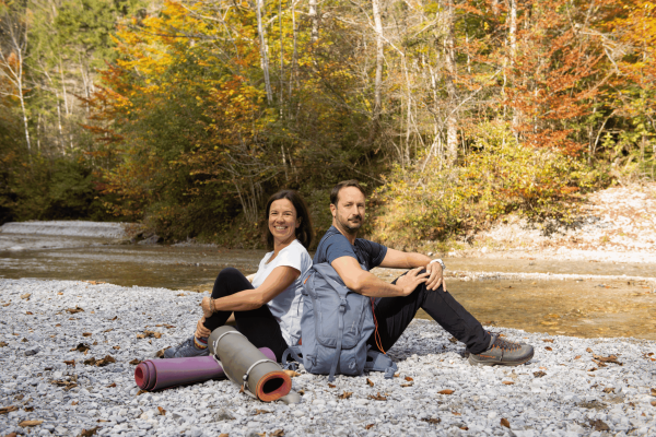 Ein Mann und eine Frau sitzen am Ufer eines Flusses in einem bewaldeten Gebiet mit bunten Herbstblättern. Die Frau lächelt, während der Mann ernst schaut. Sie sind vorbereitet für eine Wanderung mit Rucksack und Yogamatten. Es ist ein sonniger Tag im Herbst.