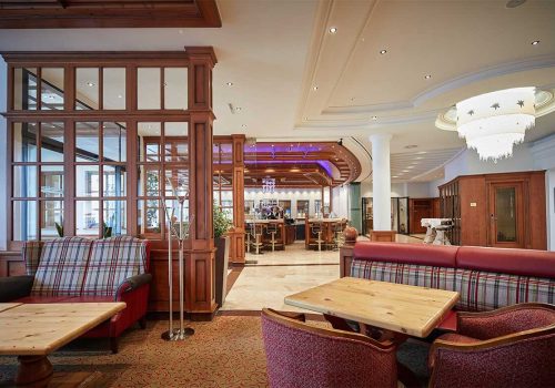 Innenraum eines eleganten Restaurants oder Hotels mit Holzmöbeln, einem Barbereich im Hintergrund, und einem Kronleuchter an der Decke.