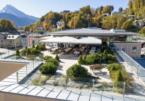 Dachterrasse eines Gebäudes mit Tischen, Stühlen, Sonnenschirmen und bepflanztem Garten, mit einer Stadt und Bergen im Hintergrund.