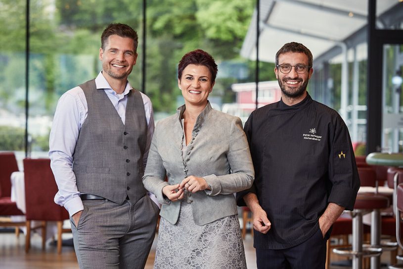 Drei gut gekleidete Menschen stehen nebeneinander in einem Restaurant oder Café, lachend und in entspannter Haltung, mit großen Fenstern im Hintergrund und Blick ins Grüne.