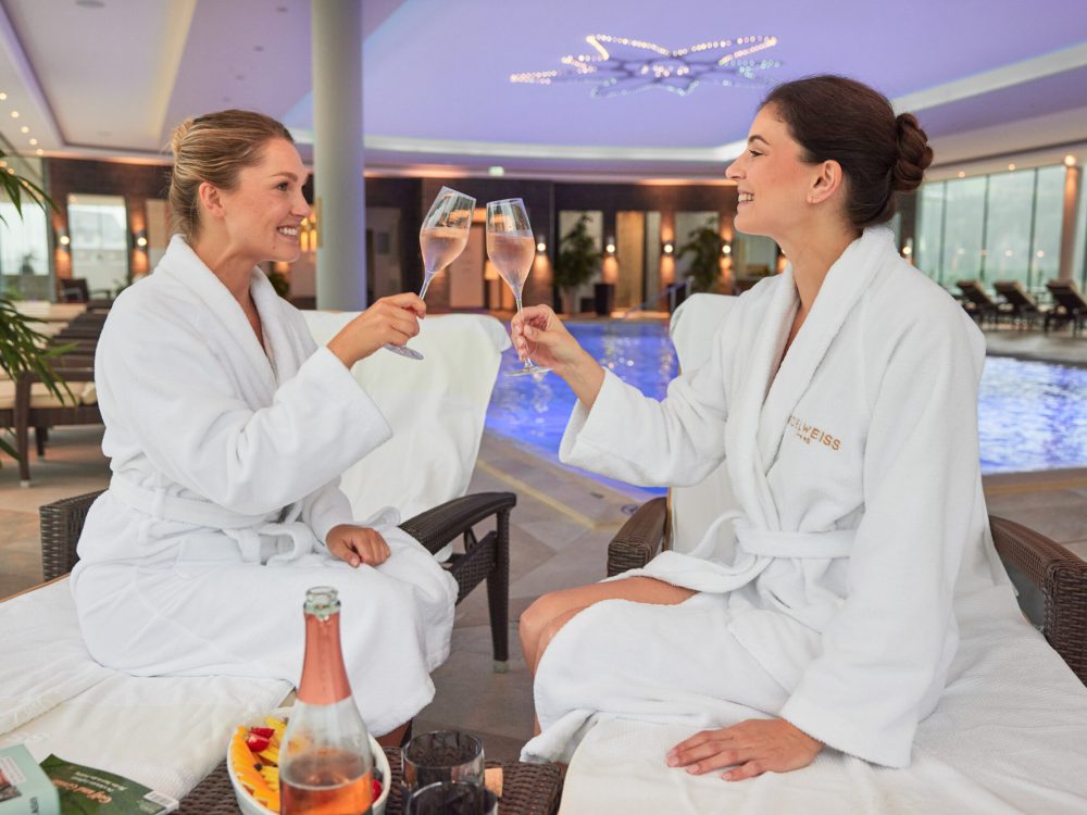 Zwei Frauen in Bademänteln sitzen auf Liegen am Pool und stoßen mit Sektgläsern an.