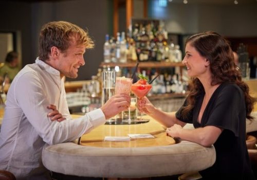 Ein Mann und eine Frau sitzen sich an einem runden Bar-Tisch gegenüber, beide lachen und stoßen mit farbenfrohen Getränken an, im Hintergrund ist die Bar mit Flaschen zu sehen.