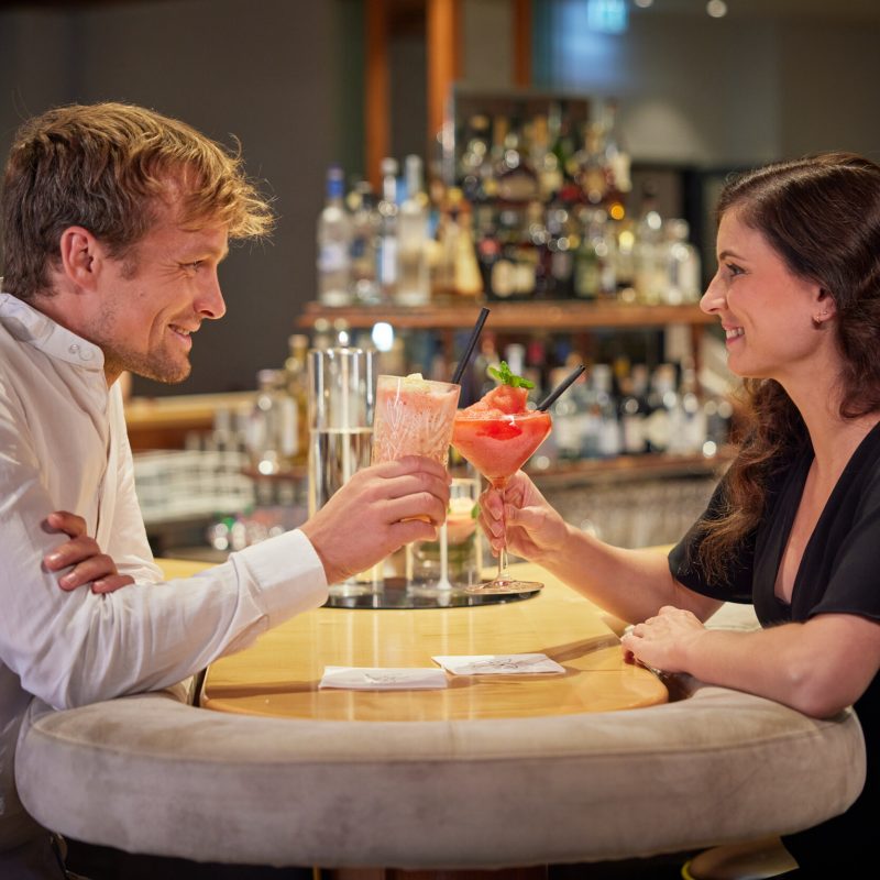 Ein Mann und eine Frau sitzen sich an einem Bar-Tisch gegenüber, beide halten bunte Cocktails in der Hand und lächeln sich an, während im Hintergrund eine Bar mit verschiedenen Flaschen zu sehen ist.