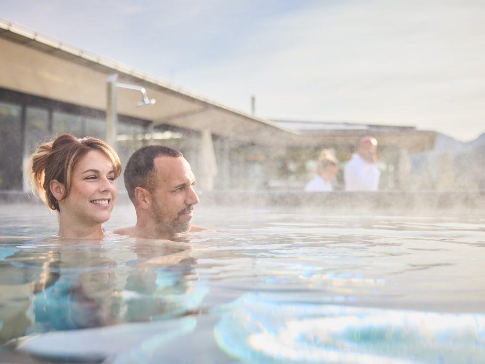 Ein Mann und eine Frau entspannen sich in einem heißen Pool im Freien, die Frau lächelt, während die Männer neben ihr sitzen, im Hintergrund sind andere Menschen und ein modernes Gebäude sichtbar.