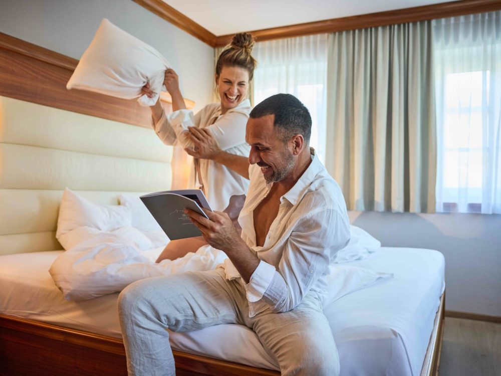 Ein Mann sitzt lachend auf der Bettkante und liest ein Buch, während eine Frau im Hintergrund mit einem Kissen auf sie wirft und ebenfalls lacht in einem hell erleuchteten Hotelzimmer.