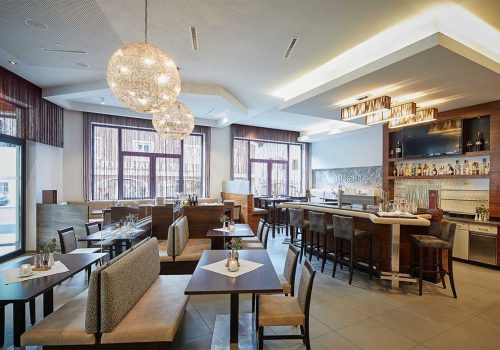 Ein gut beleuchtetes Restaurant mit modernen Dekor, mehreren Tischen mit Stühlen und einem Barbereich mit Barhockern, großen Fenstern mit Vorhängen und dekorativen Lichtleuchten an der Decke.