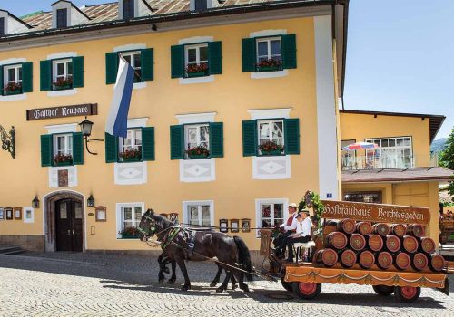 Ein Pferdegespann mit zwei Männern auf einem Kutschfahrzeug, das mit mehreren Weinfässern beladen ist, fährt an einem gelben Gebäude mit grünen Fenstergbrettern und Blumenvasen vorbei.
