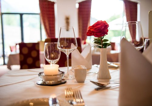 Ein gedeckter Tisch in einem Restaurant mit einer weißen Tischdecke, auf der sich eine rote Rose in einer weißen Vase, ein brennendes Teelicht, mehrere leere Weingläser, ein Messer und eine Gabel befinden.