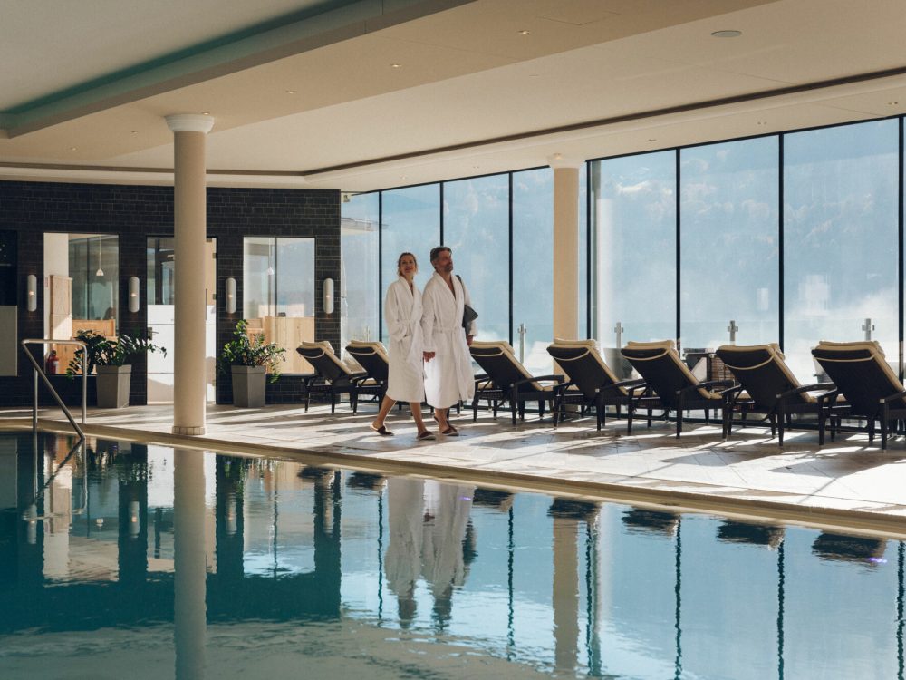Zwei Personen in weißen Bademänteln gehen an einem Innenpool vorbei, umgeben von Liegestühlen und großen Fenstern mit Blick auf Wolken und den Himmel.